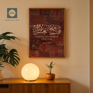 Pu&ograve; includere: Una stampa incorniciata con calligrafia araba e testo inglese: "SO, SURELY WITH HARDSHIP COMES EASE". L'opera presenta uno sfondo a motivi rossi e marroni. Una lampada rotonda e luminosa e piante in vaso sono su un mobile di legno.