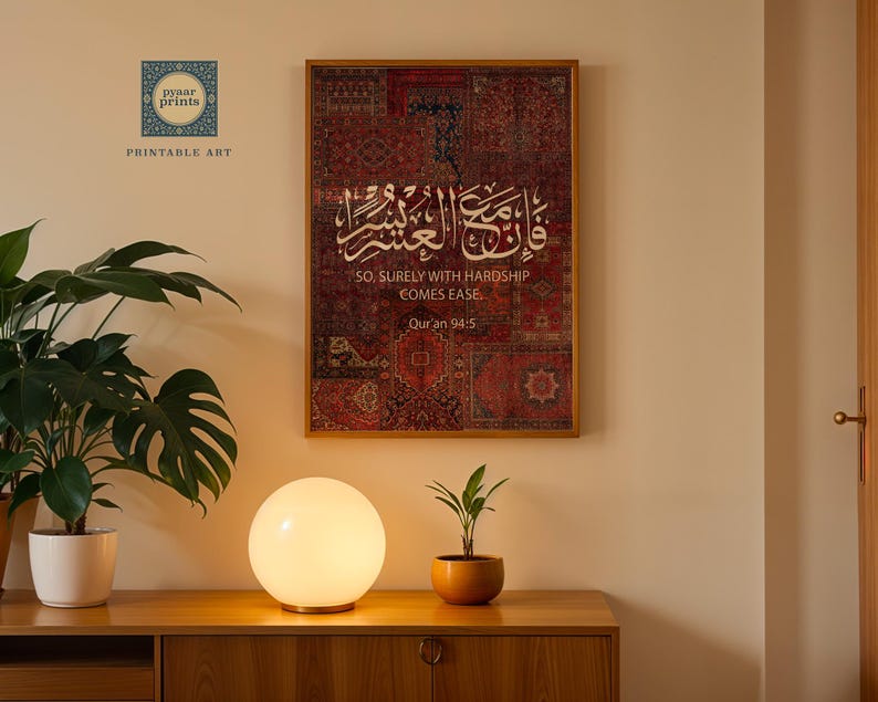 Pu&ograve; includere: Una stampa incorniciata con calligrafia araba e testo inglese: "SO, SURELY WITH HARDSHIP COMES EASE". L'opera presenta uno sfondo a motivi rossi e marroni. Una lampada rotonda e luminosa e piante in vaso sono su un mobile di legno.