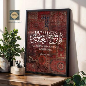 Pu&ograve; includere: Una stampa incorniciata con un motivo a patchwork di tappeti rossi e bordeaux. Calligrafia araba bianca e testo inglese: "SO, SURELY WITH HARDSHIP COMES EASE. Qur'an 94:5." La stampa &egrave; su una superficie di legno con una pianta in vaso e matite.
