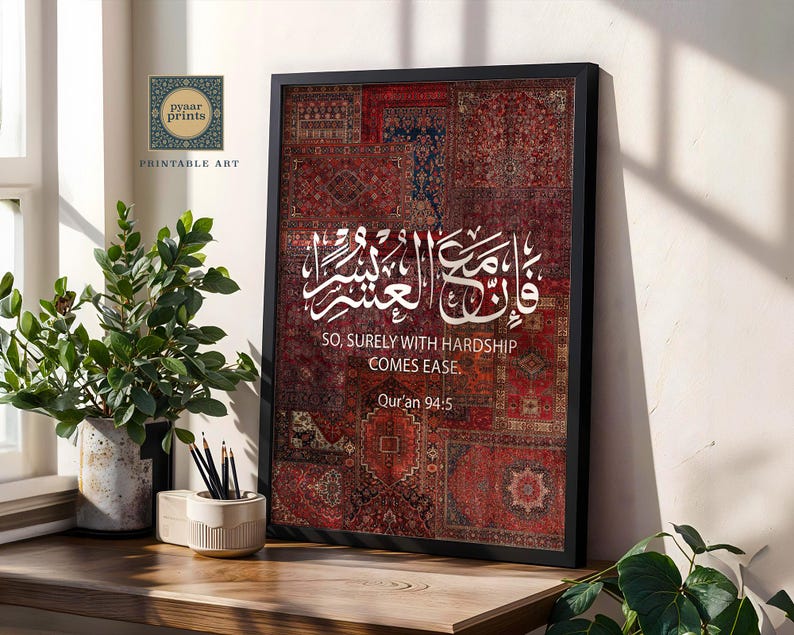 Pu&ograve; includere: Una stampa incorniciata con un motivo a patchwork di tappeti rossi e bordeaux. Calligrafia araba bianca e testo inglese: "SO, SURELY WITH HARDSHIP COMES EASE. Qur'an 94:5." La stampa &egrave; su una superficie di legno con una pianta in vaso e matite.