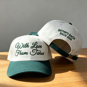 May include: Two baseball caps featuring a cream-colored crown and a dark green brim. The front cap displays the embroidered text "With Love From Tahoe" in dark green script. The back cap features the embroidered text "BRYNN'S BACH 2026" in dark green.