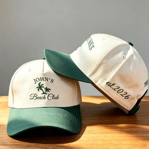 Op de afbeelding: Twee off-white baseballpetten met donkergroene kleppen. De voorste pet heeft donkergroen borduurwerk met de tekst John's Beach Club en een palmboomicoon. De zijkant van de tweede pet toont de geborduurde tekst est. 2026 in het donkergroen.