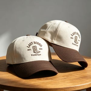 Könnte beinhalten: Zwei Baseballkappen mit cremeweißen Kronen und dunkelbraunen Schirmen. Jede Kappe zeigt eine dunkelbraune Stickerei eines Cowboystiefels mit dem Text "LAST RODEO", "EST. 2026" und dem Namen "Frederique" unter dem Stiefelmotiv.
