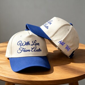 Può includere: Due cappellini da baseball con corona color crema e visiera blu. Il cappellino anteriore presenta il testo ricamato in blu "With Love, From Austin". Il secondo cappellino, posizionato dietro, mostra il testo ricamato in blu "Add Text" sul lato.