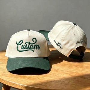 Peut inclure: Deux casquettes de baseball avec une calotte blanc cassé et une visière vert foncé. La casquette avant affiche le mot "Custom" en broderie cursive vert foncé. La deuxième casquette, placée derrière, montre le mot "custom" sur le côté.