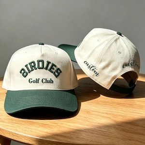 Könnte beinhalten: Zwei cremeweiße Baseballkappen mit dunkelgrünen Schirmen. Die vordere Kappe zeigt den dunkelgrün gestickten Text "BIRDIES Golf Club" in einer geschwungenen Schrift. Die Seite der hinteren Kappe zeigt das dunkelgrün gestickte Wort "custom" in Schreibschrift.