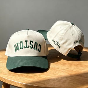 May include: Two baseball caps featuring an off-white crown and a dark green brim. The front cap displays the word CUSTOM in dark green block letters. The side of the second cap features the word custom embroidered in a dark green cursive script.