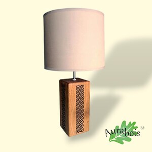 May include: A wooden table lamp with a beige lampshade. The base is a rectangular block of wood with a Celtic knot design carved into it. The lamp is lit, casting a soft glow.