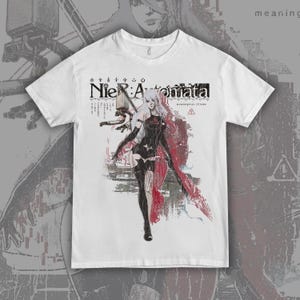 May include: White t-shirt featuring a graphic of a female character from the video game "NieR:Automata." The design includes the game's logo and Japanese text, along with a stylized illustration in black, red, and gray.
