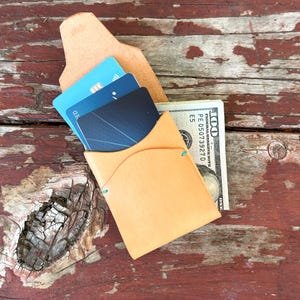 May include: A light tan leather wallet with a card slot and a cash pocket. The wallet holds several credit cards and a stack of US one hundred dollar bills. The wallet is on a weathered, reddish-brown wooden surface.