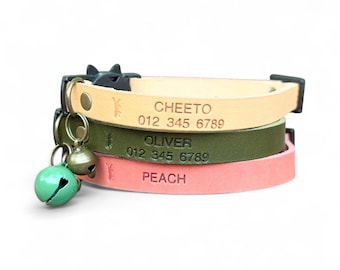May include: Stacked leather cat collars in beige, olive green, and peach. Each collar is engraved with a name and phone number. A black cat-shaped buckle and a green bell with a bronze bell are attached to the top collar.
