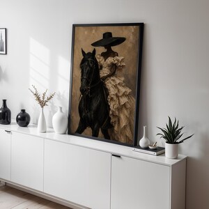 K&ouml;nnte beinhalten: Ein gerahmter Kunstdruck, der eine Person mit breitkrempigem Hut und einem cremefarbenen R&uuml;schenkleid auf einem schwarzen Pferd zeigt. Das Kunstwerk steht auf einem wei&szlig;en Sideboard neben mehreren dekorativen Vasen und einer kleinen Pflanze.
