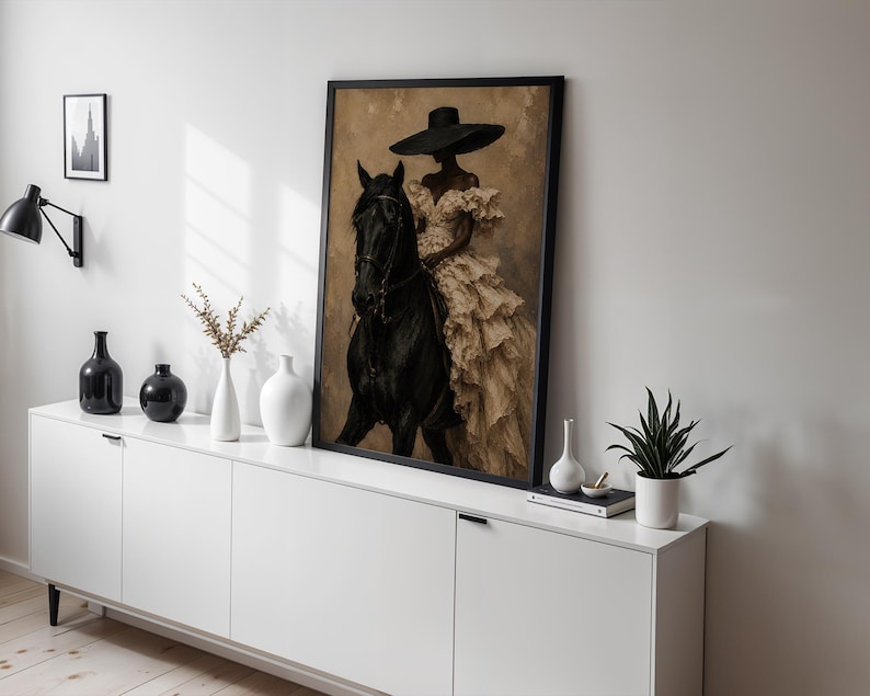 K&ouml;nnte beinhalten: Ein gerahmter Kunstdruck, der eine Person mit breitkrempigem Hut und einem cremefarbenen R&uuml;schenkleid auf einem schwarzen Pferd zeigt. Das Kunstwerk steht auf einem wei&szlig;en Sideboard neben mehreren dekorativen Vasen und einer kleinen Pflanze.