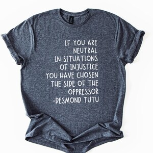 May include: Dark grey t-shirt with white text. The text reads: "IF YOU ARE NEUTRAL IN SITUATIONS OF INJUSTICE YOU HAVE CHOSEN THE SIDE OF THE OPPRESSOR -DESMOND TUTU." The shirt has a crew neck and rolled sleeves.