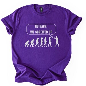Puede incluir: Una camiseta morada con un gráfico blanco que representa la evolución humana, culminando con una persona sosteniendo un teléfono inteligente. El texto "GO BACK WE SCREWED UP" está en una burbuja de diálogo encima de las figuras.