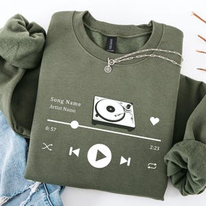 May include: An olive green sweatshirt featuring a white graphic of a record player and a music player interface. The design includes a play button, a heart, and the text "Song Name Artist Name". A silver necklace with a sun pendant is on the sweatshirt.