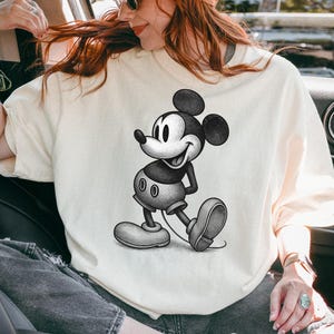 Puede incluir: Camiseta color crema con una ilustraci&oacute;n en blanco y negro de Mickey Mouse. El personaje de dibujos animados se representa en una pose cl&aacute;sica, sonriendo. La camiseta tiene un corte casual y oversize.