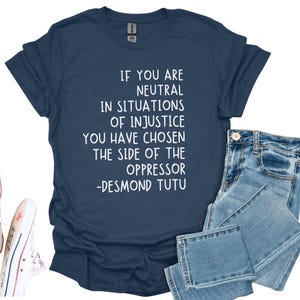 May include: Navy blue t-shirt with the quote "IF YOU ARE NEUTRAL IN SITUATIONS OF INJUSTICE YOU HAVE CHOSEN THE SIDE OF THE OPPRESSOR -DESMOND TUTU" in white. The shirt is paired with blue jeans and white Converse trainers.