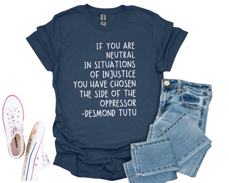 May include: Navy blue t-shirt with the quote "IF YOU ARE NEUTRAL IN SITUATIONS OF INJUSTICE YOU HAVE CHOSEN THE SIDE OF THE OPPRESSOR -DESMOND TUTU" in white. The shirt is paired with blue jeans and white Converse trainers.
