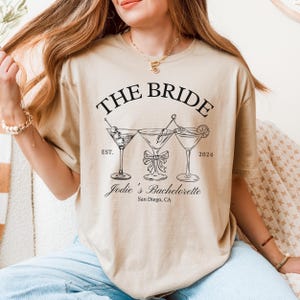 May include: Beige t-shirt with the text "THE BRIDE" above three martini glasses. Below the glasses is "Jodie's Bachelorette San Diego, CA". The shirt has a relaxed fit.