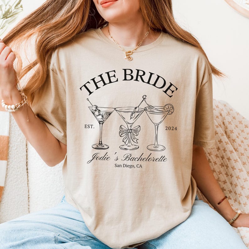 May include: Beige t-shirt with the text "THE BRIDE" above three martini glasses. Below the glasses is "Jodie's Bachelorette San Diego, CA". The shirt has a relaxed fit.