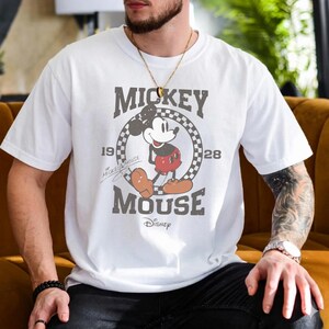 Puede incluir: Una camiseta blanca de manga corta con un gr&aacute;fico de Mickey Mouse dentro de un c&iacute;rculo a cuadros. El texto dice Mickey Mouse, 1928 y Disney. El dise&ntilde;o incluye un estampado de estilo vintage desgastado con colores rojo, negro y blanco.
