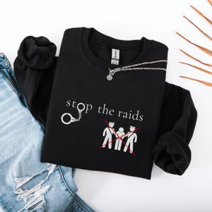 May include: Black sweatshirt with the words "stop the raids" and a graphic of handcuffs and crossed-out figures. A silver necklace is draped across the sweatshirt. The sweatshirt is displayed with a pair of blue jeans.