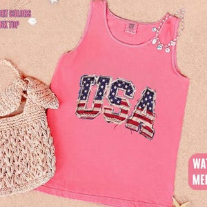 May include: A watermelon pink tank top with the word "USA" in a patriotic design. The tank top is paired with a woven beige handbag and a necklace. The words "Comfort Colors Tank Top" and "Watermelon" are also visible.