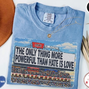 May include: A light blue Comfort Colors t-shirt with a graphic that reads "The Only Thing More Powerful Than Hate Is Love" over a stadium scene. The shirt has a crew neck and short sleeves. A gold necklace is draped across the shirt.