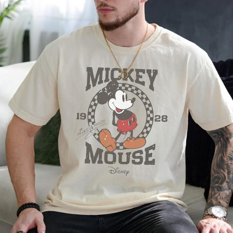 Puede incluir: Una camiseta de manga corta color crema con un gr&aacute;fico de estilo vintage de Mickey Mouse dentro de un c&iacute;rculo a cuadros. El texto dice Mickey Mouse, 1928 y Disney. El dise&ntilde;o incluye una firma y una apariencia desgastada y envejecida.