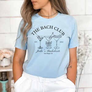 May include: Light blue t-shirt with a black graphic of three martini glasses and the text "THE BACH CLUB" above "Jodie's Bachelorette" and "San Diego, CA". The shirt has short sleeves.