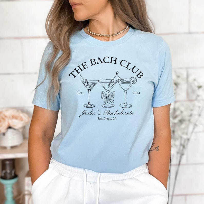 May include: Light blue t-shirt with a black graphic of three martini glasses and the text "THE BACH CLUB" above "Jodie's Bachelorette" and "San Diego, CA". The shirt has short sleeves.