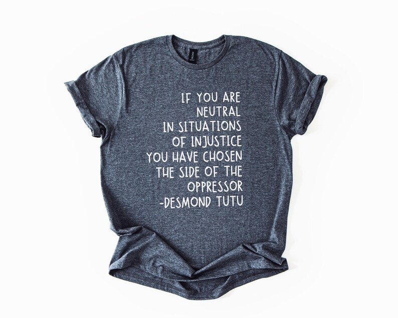 May include: Dark grey t-shirt with white text. The text reads: "IF YOU ARE NEUTRAL IN SITUATIONS OF INJUSTICE YOU HAVE CHOSEN THE SIDE OF THE OPPRESSOR -DESMOND TUTU." The shirt has a crew neck and rolled sleeves.
