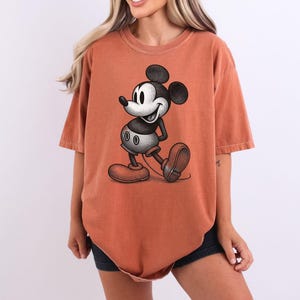 Puede incluir: Una camiseta de color naranja rojizo con una ilustraci&oacute;n en blanco y negro de Mickey Mouse. El personaje de dibujos animados se muestra caminando, con sus grandes orejas y zapatos rojos. La camiseta tiene mangas cortas y un corte holgado.