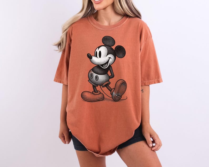 Puede incluir: Una camiseta de color naranja rojizo con una ilustraci&oacute;n en blanco y negro de Mickey Mouse. El personaje de dibujos animados se muestra caminando, con sus grandes orejas y zapatos rojos. La camiseta tiene mangas cortas y un corte holgado.