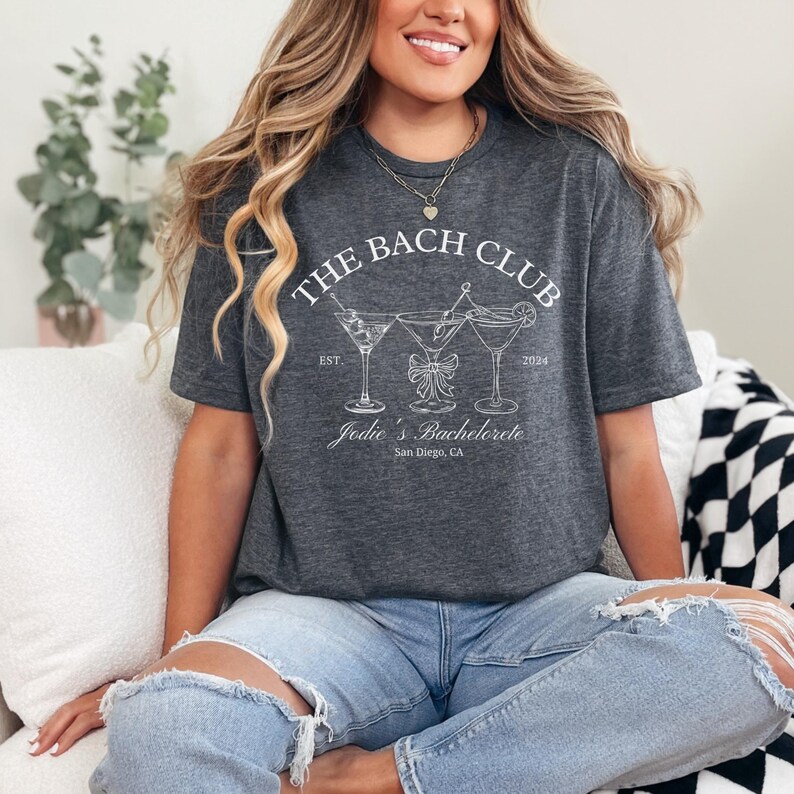 May include: Dark gray t-shirt with the text "THE BACH CLUB" above three martini glasses and the text "Jodie's Bachelorette San Diego, CA". The shirt is a casual style.