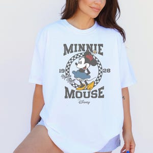 Puede incluir: Una camiseta blanca de manga corta con un gr&aacute;fico de Minnie Mouse dentro de un c&iacute;rculo a cuadros blanco y negro. El texto dice MINNIE MOUSE, 1928 y Disney. Minnie Mouse lleva una falda azul, zapatos amarillos y un lazo rojo en el pelo.