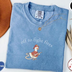 Może przedstawiać: Jasnoniebieska koszulka Comfort Colors z napisem "off to fight fires" i grafiką strażaka-kaczki z kreskówki. Koszulka ma okrągły dekolt i krótkie rękawy. Złoty naszyjnik jest przerzucony przez koszulkę.