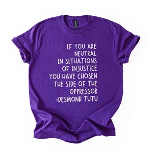 May include: A purple t-shirt with white text. The text reads: "IF YOU ARE NEUTRAL IN SITUATIONS OF INJUSTICE YOU HAVE CHOSEN THE SIDE OF THE OPPRESSOR -DESMOND TUTU."