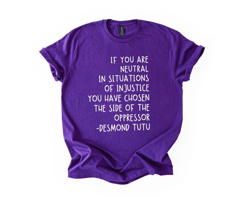 May include: A purple t-shirt with white text. The text reads: "IF YOU ARE NEUTRAL IN SITUATIONS OF INJUSTICE YOU HAVE CHOSEN THE SIDE OF THE OPPRESSOR -DESMOND TUTU."