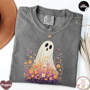 Puede incluir: Una camiseta gris Comfort Colors con un diseño de fantasma y un arreglo floral. El fantasma tiene ojos y boca negros. Las flores son en tonos naranja, amarillo, rosa y morado. La camiseta también tiene un collar plateado.