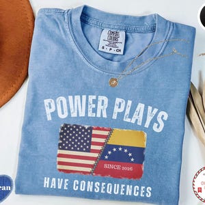 Puede incluir: Una camiseta azul claro con las palabras "POWER PLAYS HAVE CONSEQUENCES" en letras blancas. La camiseta presenta un gráfico de las banderas estadounidense y venezolana. Un collar de oro está drapeado sobre la camiseta.