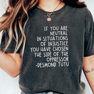 May include: Dark grey t-shirt featuring the quote "IF YOU ARE NEUTRAL IN SITUATIONS OF INJUSTICE YOU HAVE CHOSEN THE SIDE OF THE OPPRESSOR -DESMOND TUTU" in white text. The shirt has a crew neck.