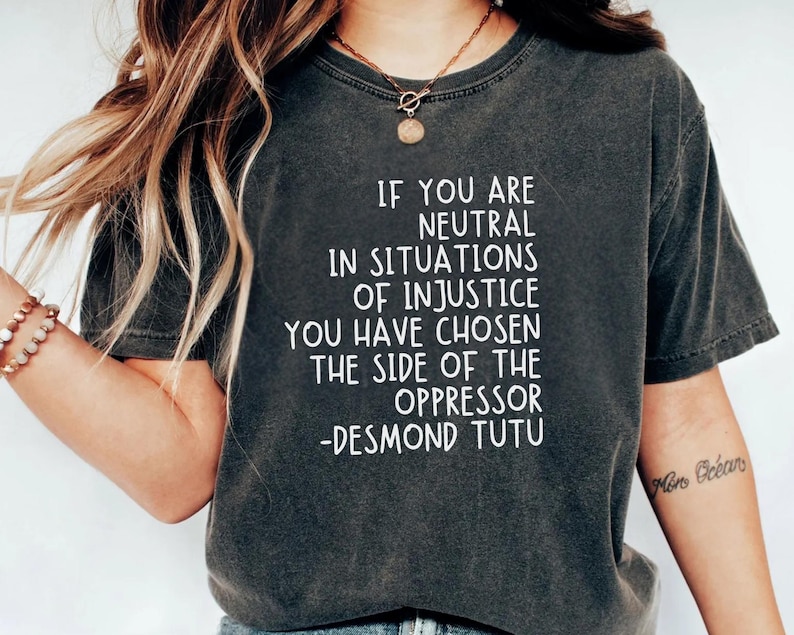 May include: Dark grey t-shirt featuring the quote "IF YOU ARE NEUTRAL IN SITUATIONS OF INJUSTICE YOU HAVE CHOSEN THE SIDE OF THE OPPRESSOR -DESMOND TUTU" in white text. The shirt has a crew neck.