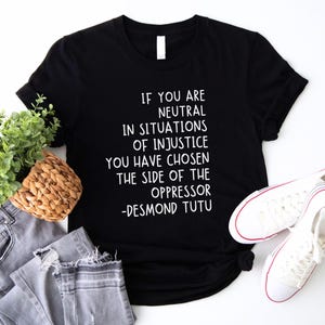 May include: Black t-shirt with a quote by Desmond Tutu: "If you are neutral in situations of injustice, you have chosen the side of the oppressor." The image includes grey jeans, white trainers, and a small plant in a woven basket.