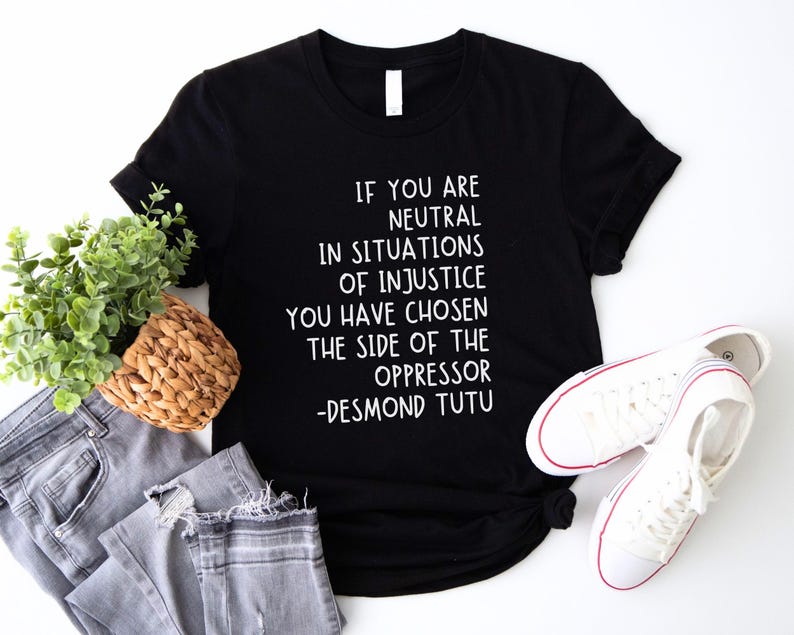 May include: Black t-shirt with a quote by Desmond Tutu: "If you are neutral in situations of injustice, you have chosen the side of the oppressor." The image includes grey jeans, white trainers, and a small plant in a woven basket.