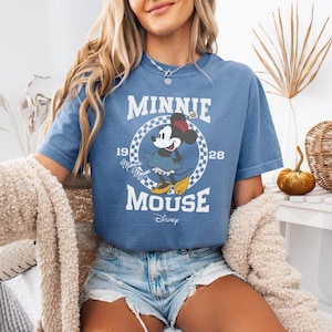 Puede incluir: Una camiseta azul de manga corta con un gr&aacute;fico de Minnie Mouse dentro de un c&iacute;rculo a cuadros. El texto dice MINNIE MOUSE, 1928 y Disney. El dise&ntilde;o incluye una peque&ntilde;a flor, la firma Minnie Mouse XOXO y una est&eacute;tica vintage.