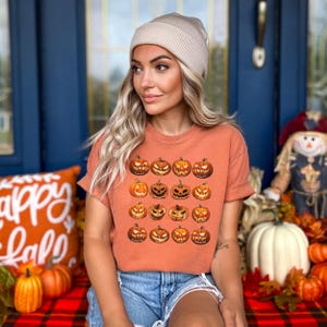 以下が含まれることがあります： オレンジ色のTシャツ。16個の彫刻されたカボチャの顔がグリッド状に並んでいます。さまざまな表情があり、オレンジと茶色の色合いです。デニムショーツとベージュの帽子を着用しています。背景には秋の装飾があります。