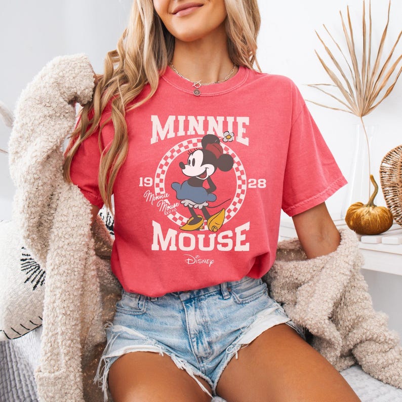 Puede incluir: Una camiseta de manga corta color rojo coral con un gr&aacute;fico de Minnie Mouse dentro de un c&iacute;rculo a cuadros blanco y rojo. El texto dice Minnie Mouse 1928 con el logotipo de Disney debajo. La camiseta se lleva con pantalones cortos de mezclilla azul claro y un c&aacute;rdigan beige.