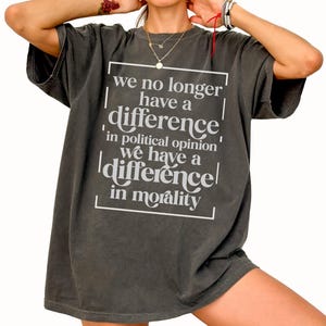 May include: Dark gray oversized t-shirt with white text. The text reads "we no longer have a difference in political opinion we have a difference in morality" within a white rectangular border. The shirt has short sleeves.